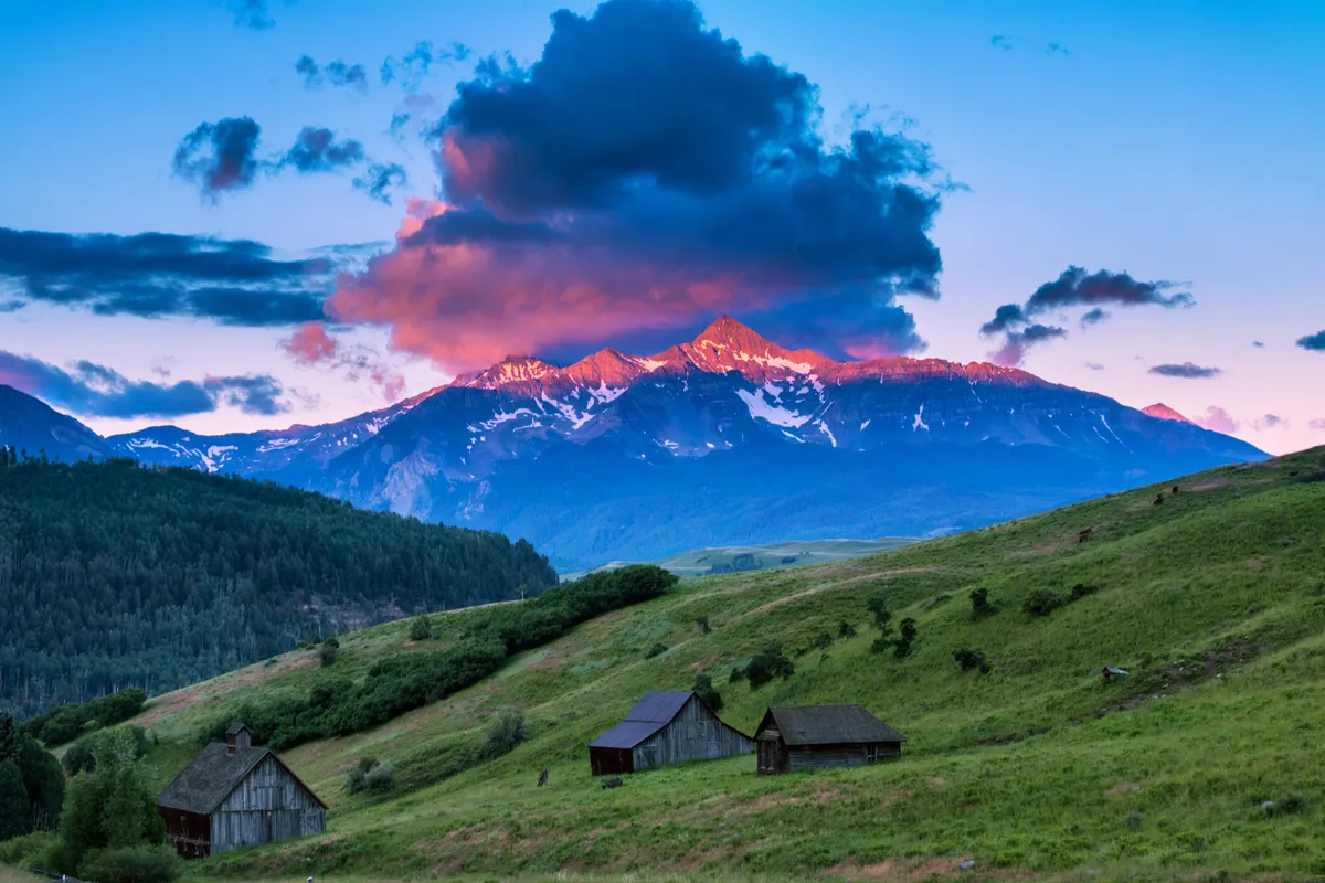 This image features a sweeping view of majestic mountains under colorful sunset clouds, with wooden cabins nestled among lush green hills. The natural scenery suggests outdoor adventure and the beauty of Telluride's environment, inviting guests to explore rather than stay indoors.