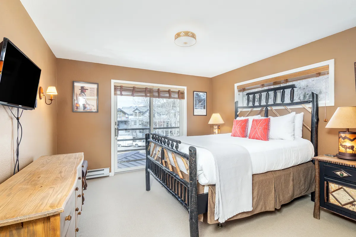 The image showcases a cozy interior with a plush king-sized bed positioned near a large window, allowing natural light to fill the room. The décor features a wooden dresser, tasteful artwork, and warm lighting from bedside lamps, creating an inviting and comfortable ambiance.