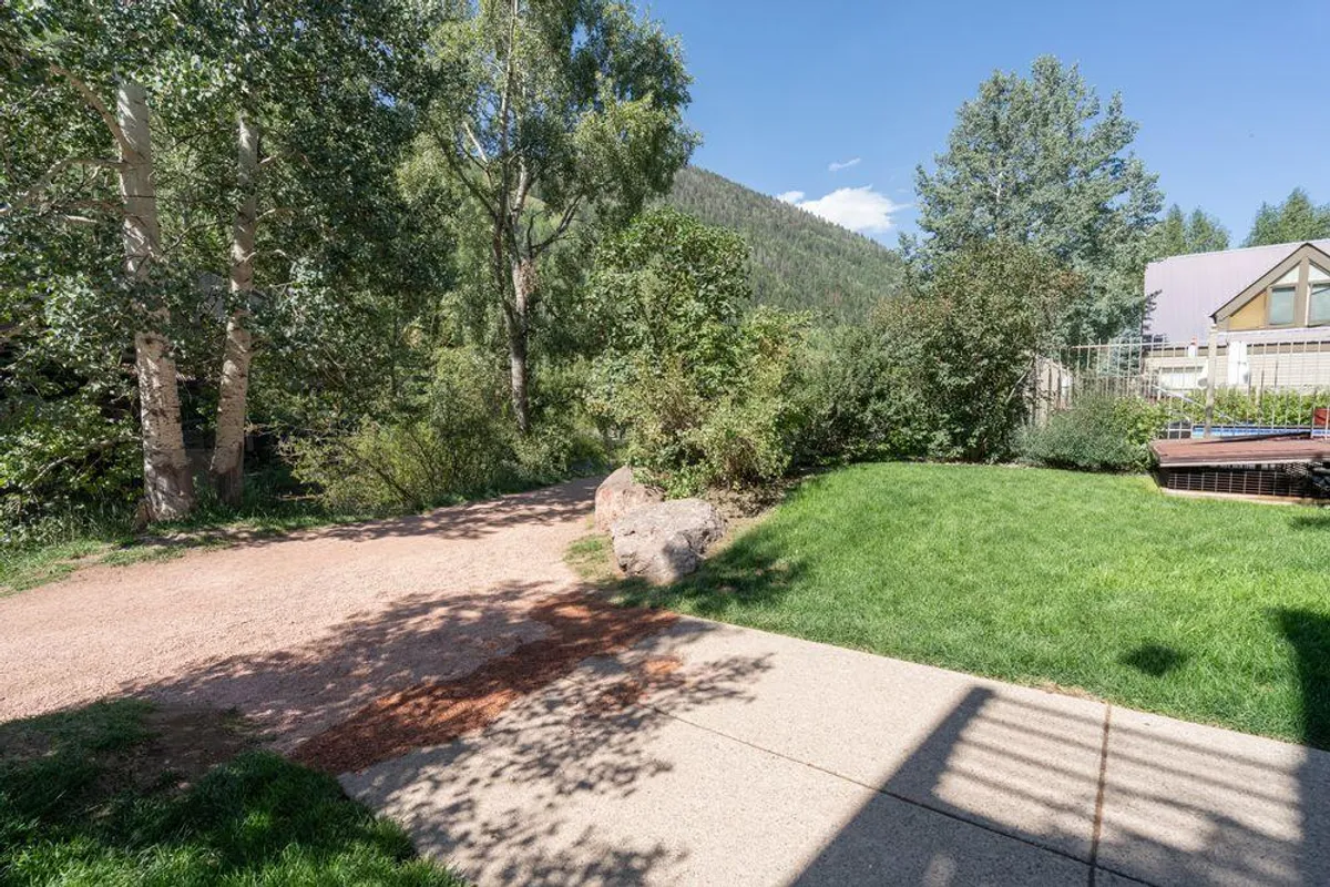 A tranquil exterior scene featuring a winding pathway bordered by lush greenery, with a spacious deck area overlooking a gentle river. The backdrop is filled with mountains, and the setting exudes a sense of peace. The environment invites walking, relaxing, and enjoying nature, with a deck perfect for sipping coffee or hosting a riverside picnic.