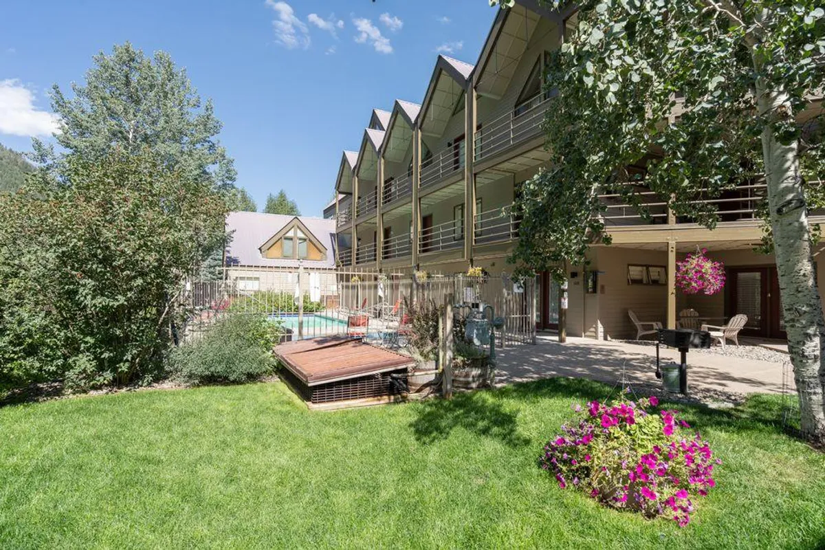 A cheerful outdoor pool scene surrounded by balconies and flower beds, with a lush green lawn and seating areas. The pool is the central focus, inviting for a swim or relaxation on a sunny day. Several balconies overlook the pool, suggesting easy access from the building and a strong sense of community.