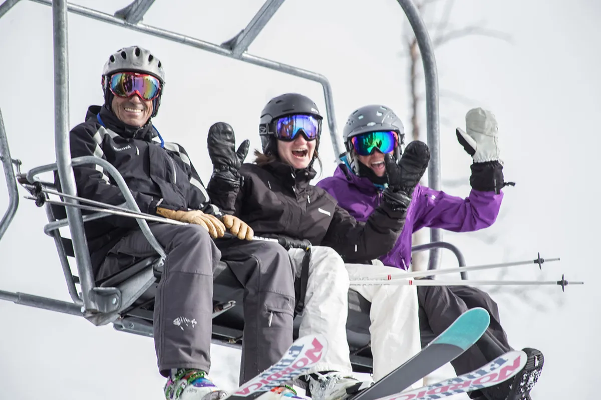 Three skiers ride a chairlift above a snowy landscape, their ski equipment visible as they share a joyful moment. The expansive view below highlights the vastness of the mountain terrain and the exhilarating experience of skiing in Telluride. The image radiates a sense of fun and camaraderie associated with mountain adventure.