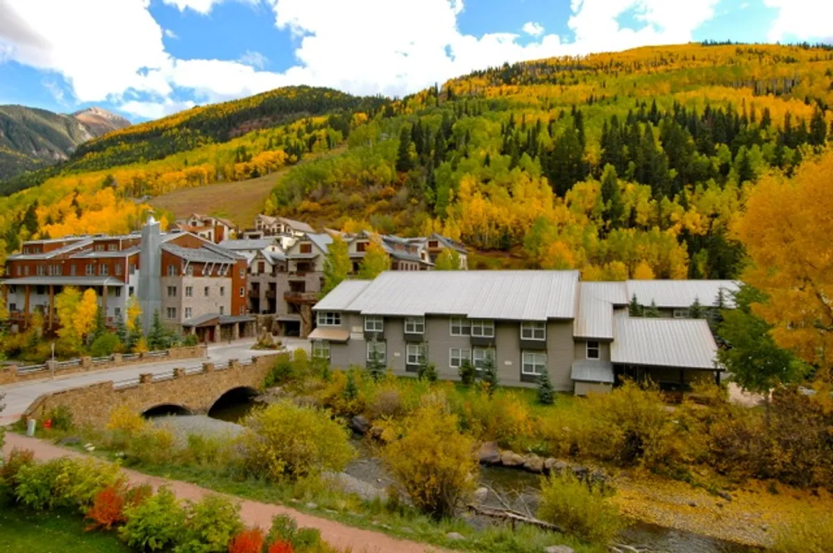 A tranquil riverside scene showing colorful autumn foliage, a gently flowing river, and buildings along the riverbank. The natural setting emphasizes the proximity to outdoor trails and the beauty of the surrounding environment.