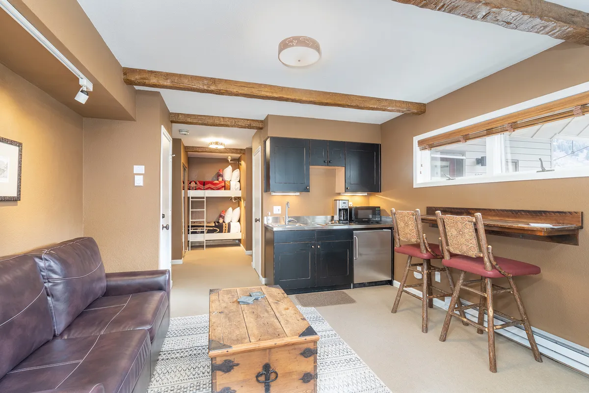 A bright, welcoming living area featuring a leather sofa, wooden coffee table, and kitchenette with bar stools. The space is ideal for relaxing after a day outdoors, socializing, or enjoying movie nights—activities highlighted in the section content.
