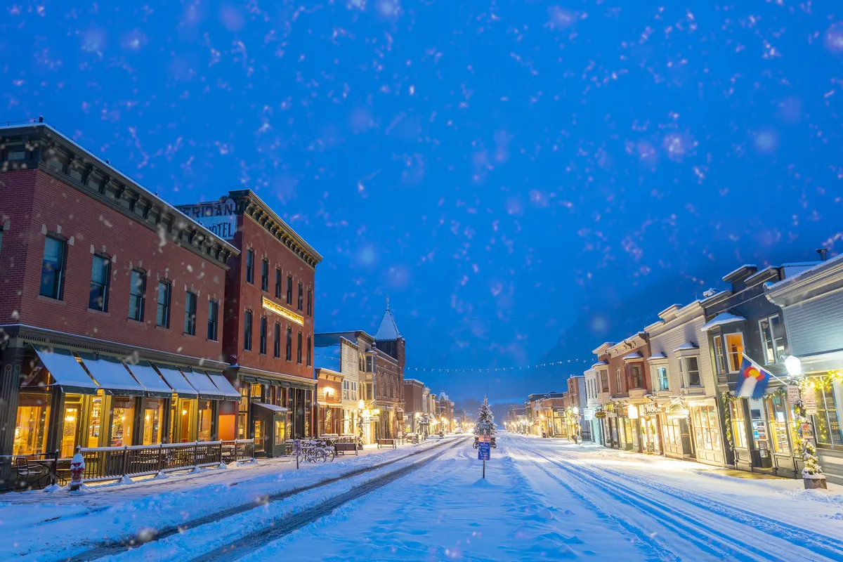 A snow-covered street in downtown Telluride adorned with decorated storefronts, twinkling holiday lights, and a central tree. Locals and visitors alike stroll the festive avenue, pausing to window-shop or savor hot drinks. The scene glows with a warm, cozy energy, perfect for evening exploration.