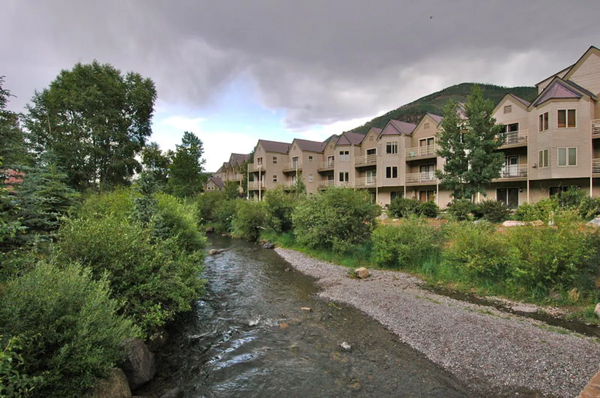 A peaceful view of a stream running beside an apartment complex, framed by lush greenery and walking paths. The complex is nestled in nature, inviting residents to enjoy serene mornings by the water and explore the outdoors.