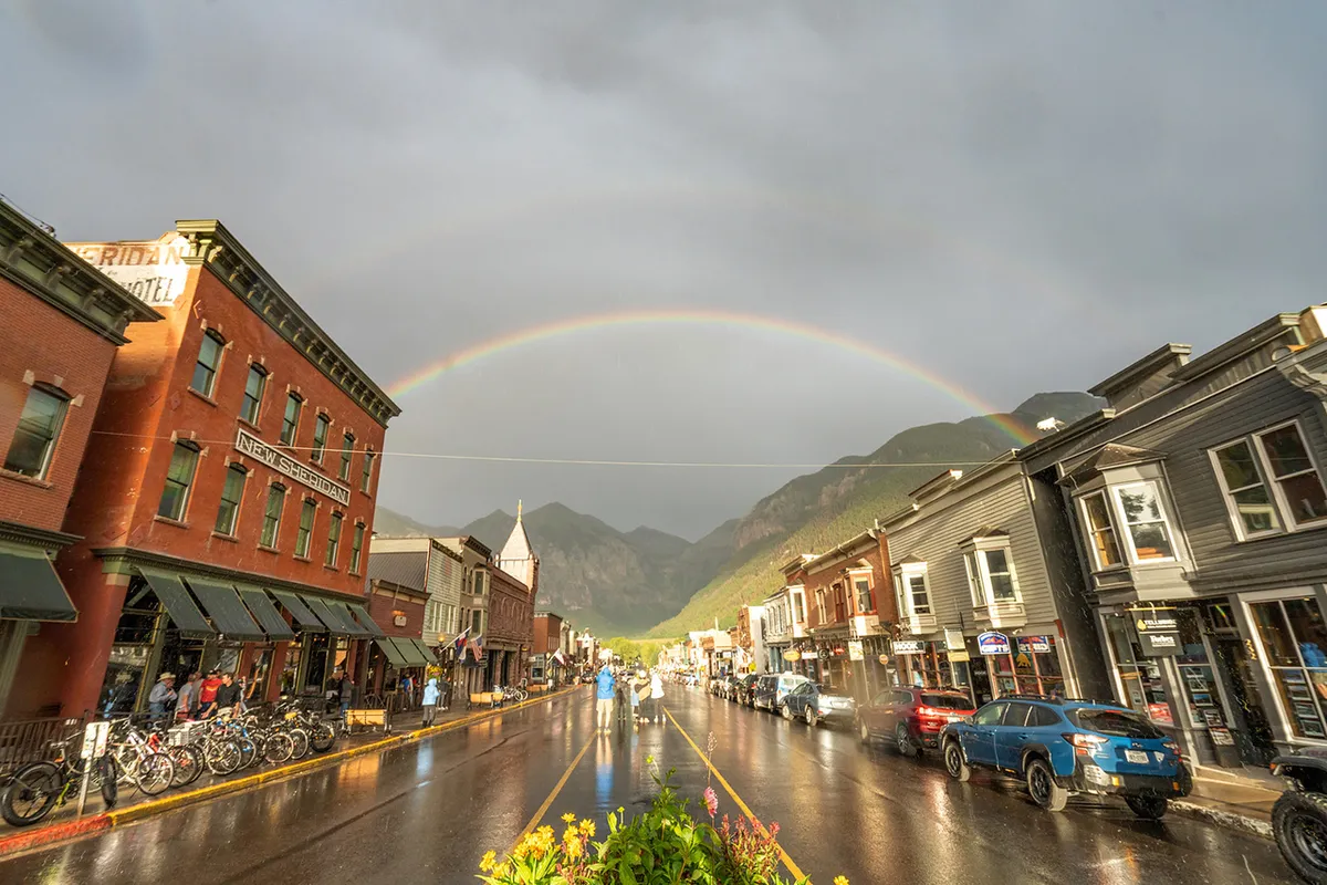 A vibrant street scene in downtown Telluride featuring historic buildings lining a wet street, with a rainbow stretching overhead. People stroll along the sidewalks, some riding bicycles, while others pause at shop entrances or enjoy outdoor café seating. The unique charm of the town is highlighted by the mixture of colorful storefronts and lively street activity.
