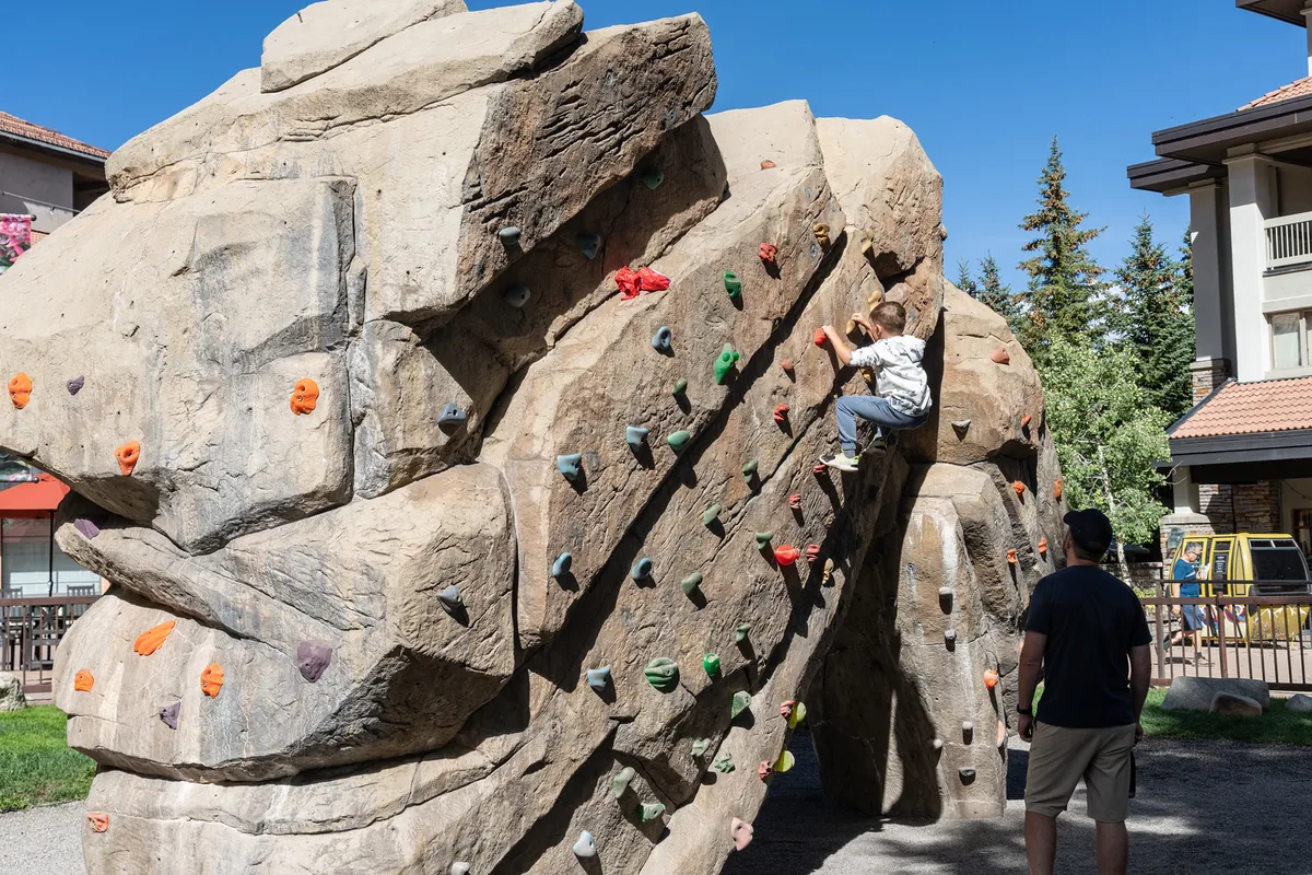 A child is actively climbing on an outdoor climbing wall while an adult stands nearby, observing and encouraging. The surrounding area is open, with greenery that suggests a park-like setting perfect for families and outdoor activities.