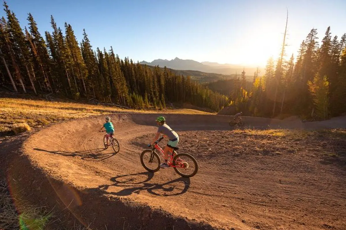 A group of mountain bikers rides along a scenic mountain trail at sunset, surrounded by pine trees and a dramatic mountain range. The image is lively, capturing the freedom and exhilaration of summer mountain biking and hiking—core activities accessible from the property.